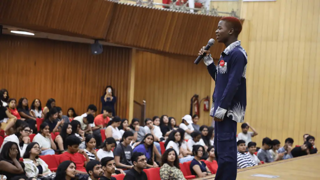 Person with a microphone speaks to an engaged audience seated in a wooden auditorium.