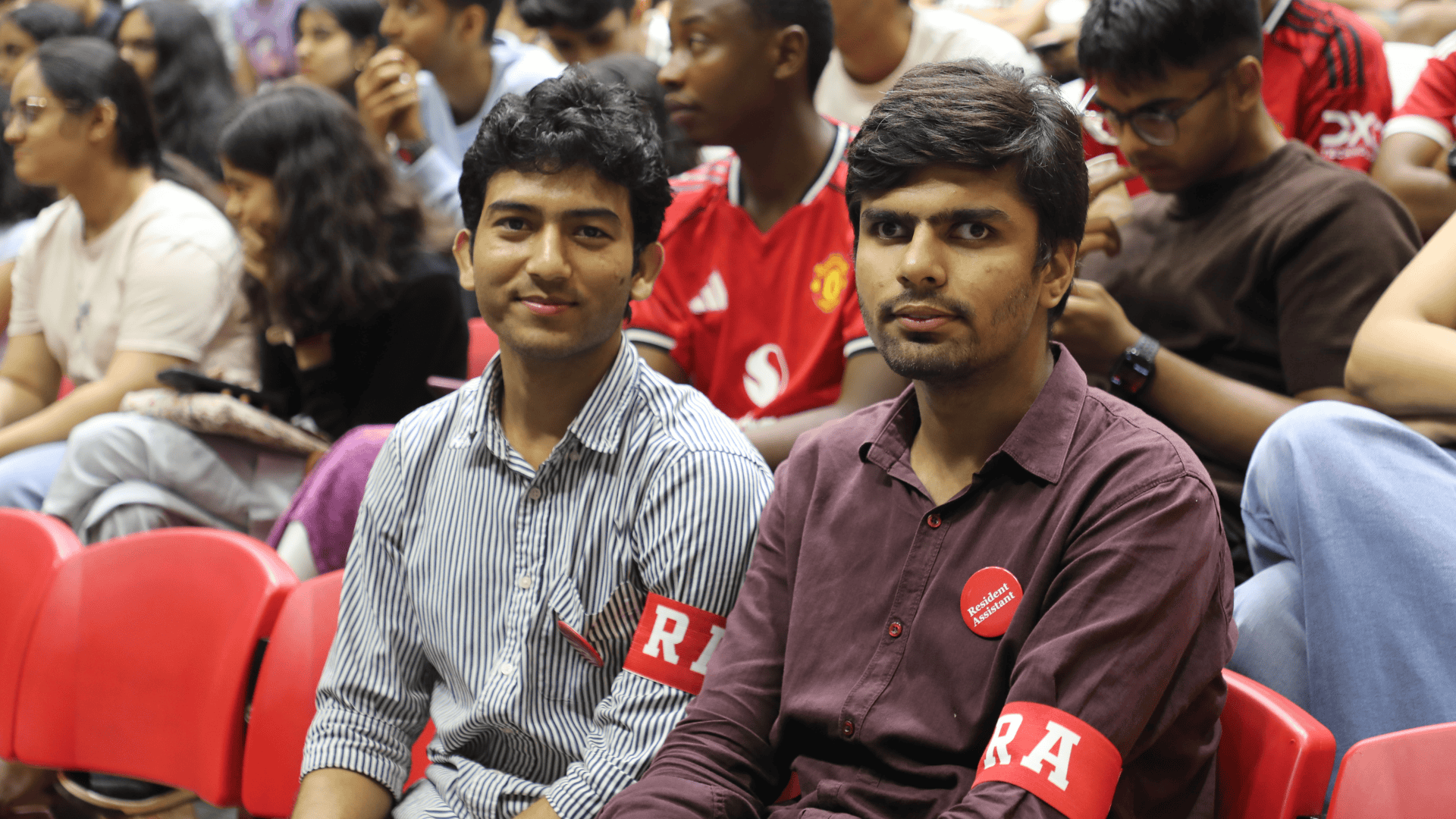 Two men wearing "RA" armbands sit in a crowd, with others seated in red chairs behind them.