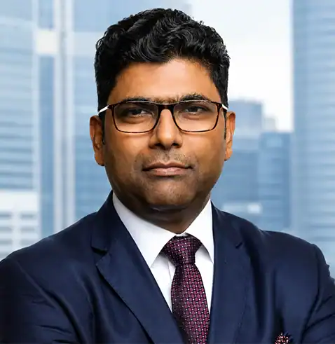 Man in a navy suit, white shirt, and maroon tie, wearing glasses, with a cityscape background.