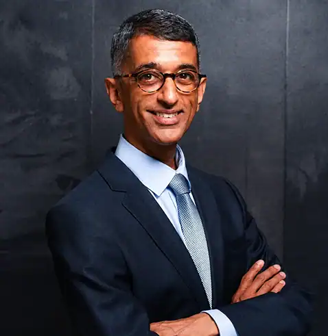 Man in a dark suit, light blue tie, and glasses, smiling with arms crossed against a dark background.