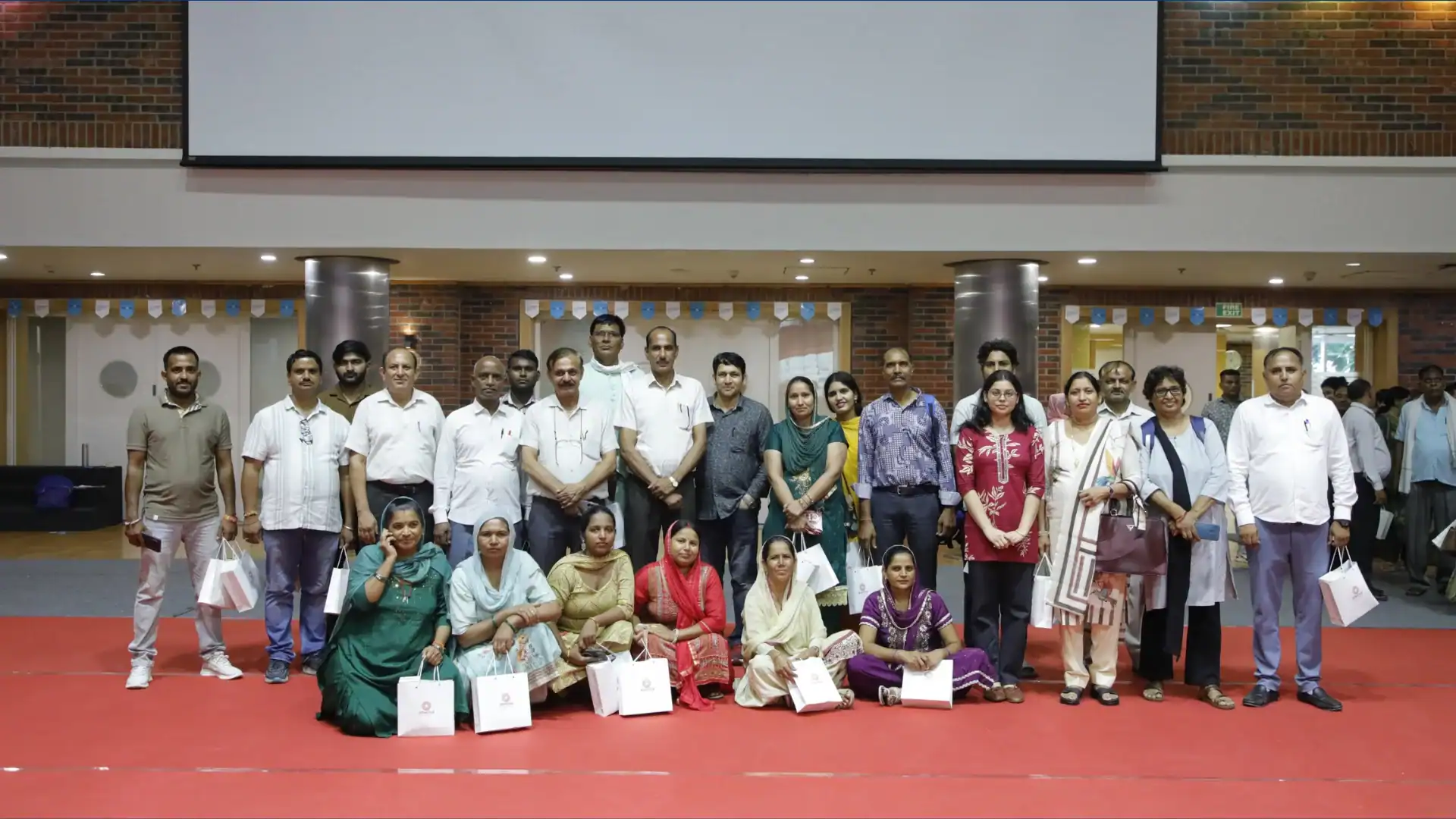 A group of men and women posing indoors on a red carpet, some holding white gift bags.