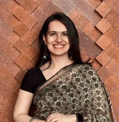 Smiling woman in a patterned saree with black blouse, standing against a textured brick wall.
