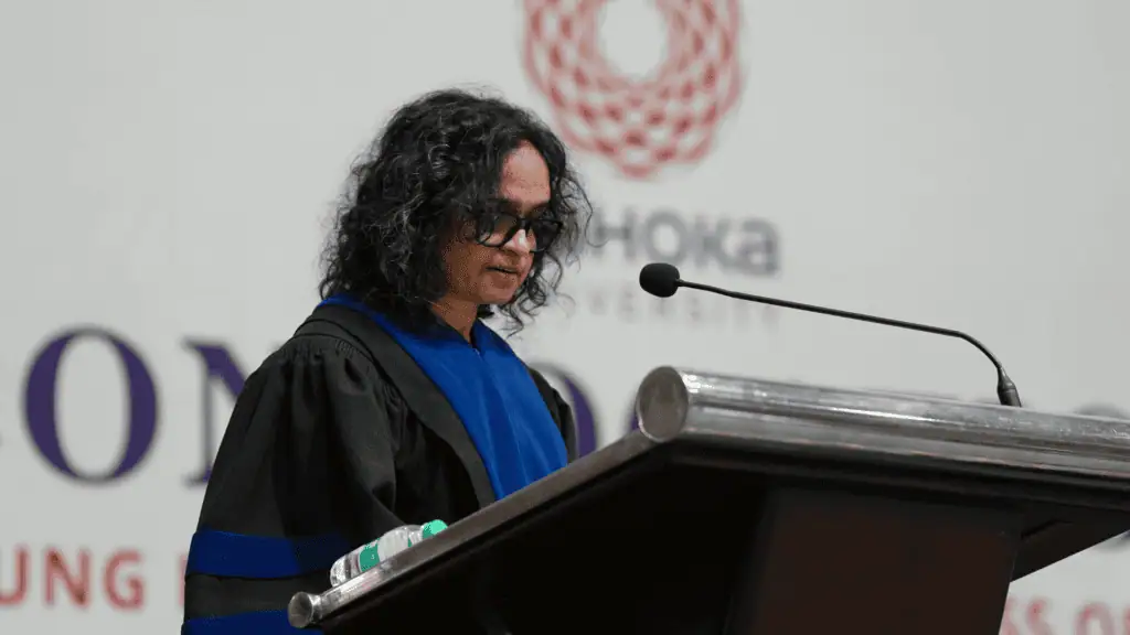 Person in academic robes speaking at a podium with a microphone, university logo in the background.
