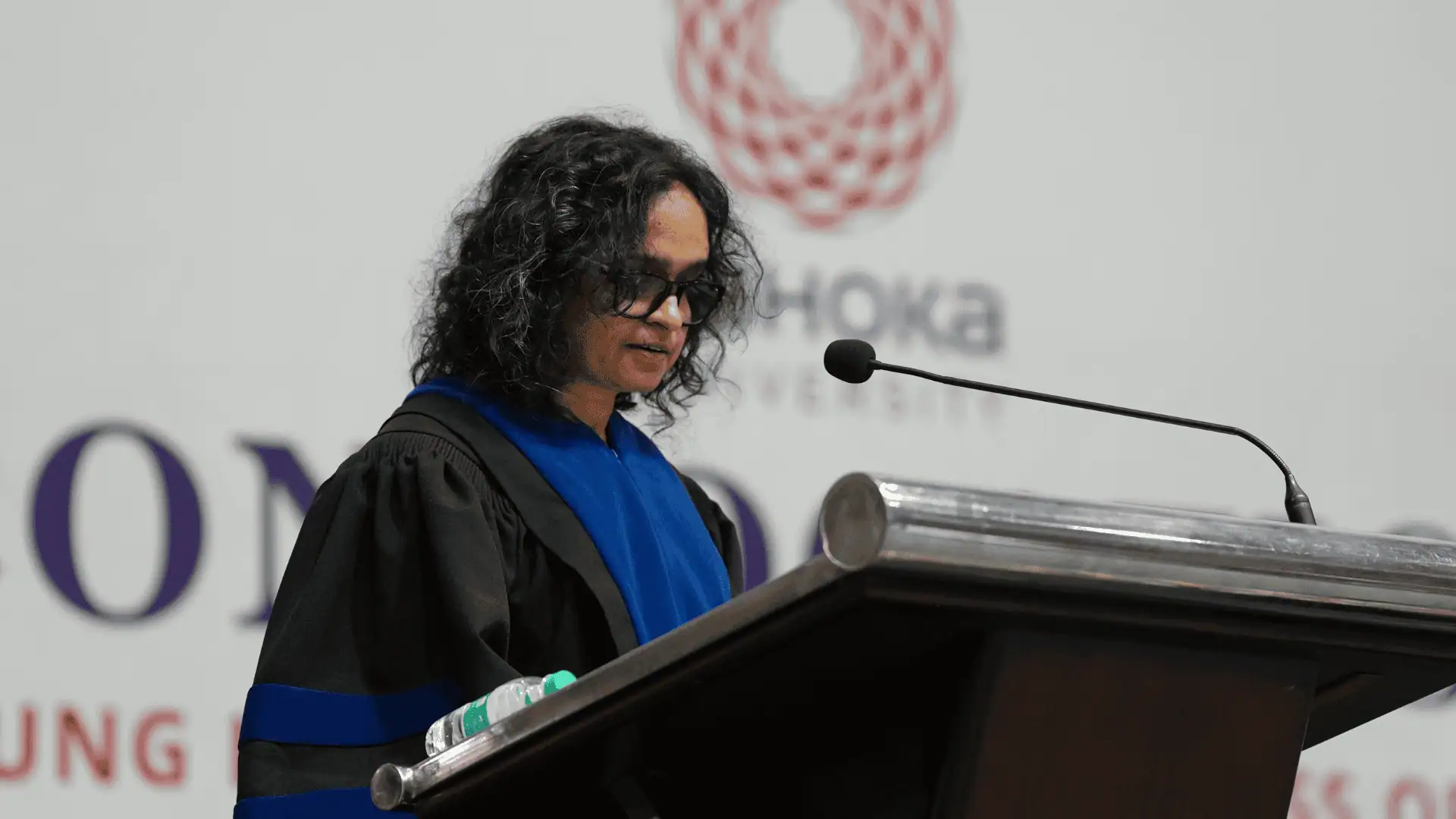 Person in academic robes speaking at a podium with a microphone, university logo in the background.
