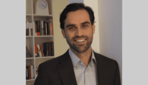 Smiling man in a suit with a bookshelf and clock in the background.
