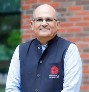 Smiling man in glasses wearing a black vest with Ashoka University logo, standing near a brick wall.